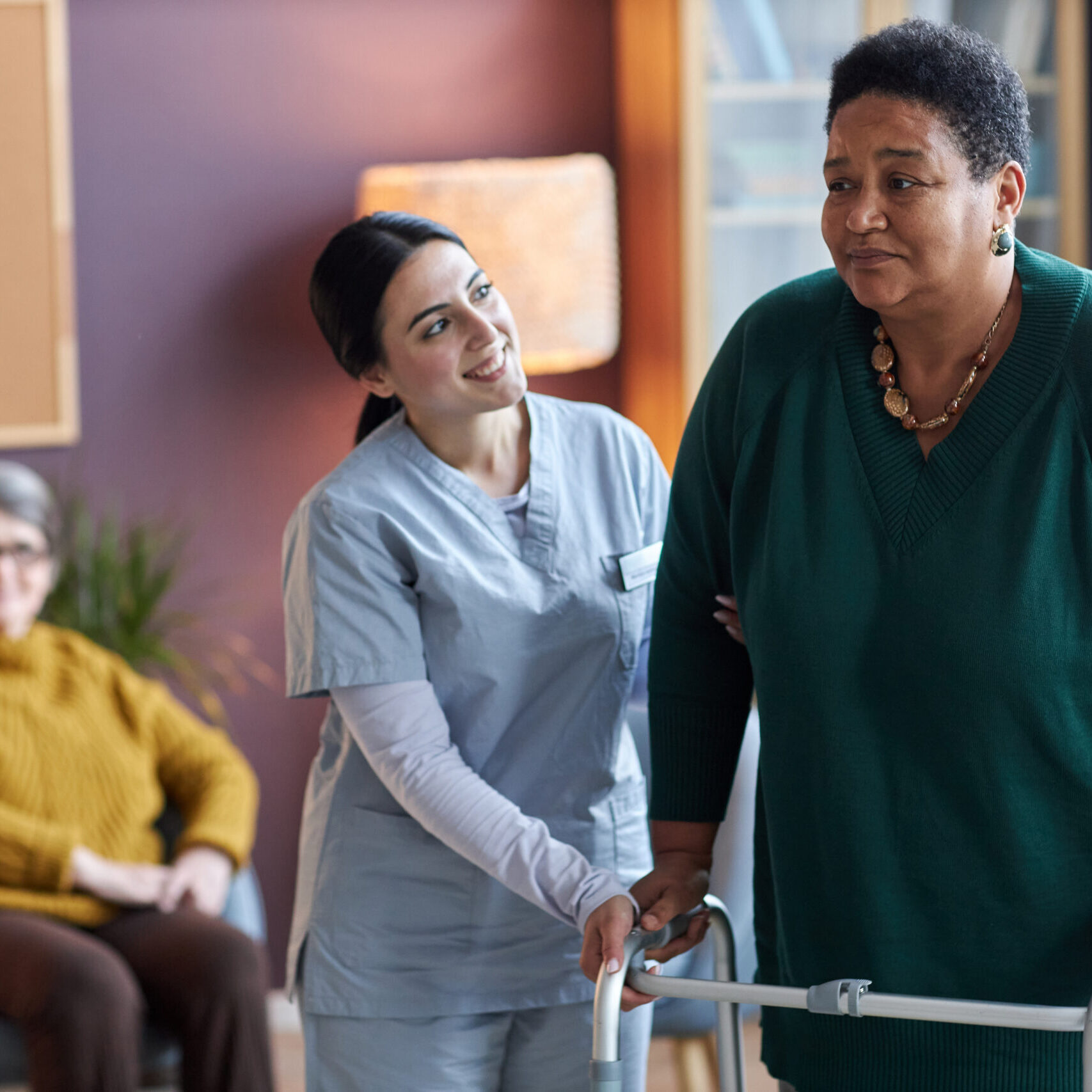 Portrait of smiling young nurse helping black senior woman using mobility walker in retirement home, copy space